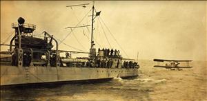 A large warship tows a single propellor plane through choppy seas. Sailors lean over the railing of the ship. 